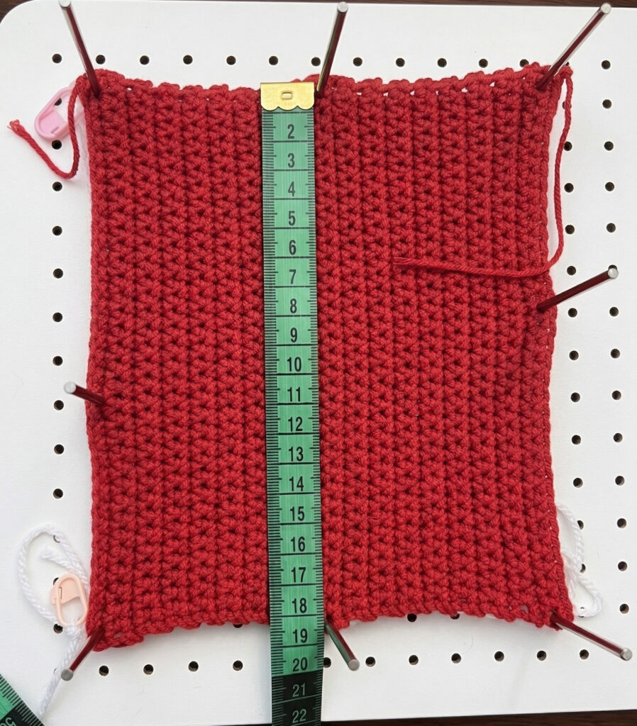 Amostra vermelha de crochê fixada em placa de blocagem, sendo medida com uma fita métrica verde marcando exatamente 18 centímetros. Etapa de planejamento para o casaco de Pixel Art em Crochê.