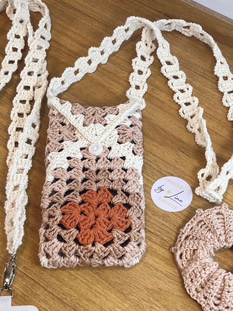 Bolsa para celular de crochê artesanal com estampa granny square em tons neutros de bege e marrom, com botão branco e alça longa.
