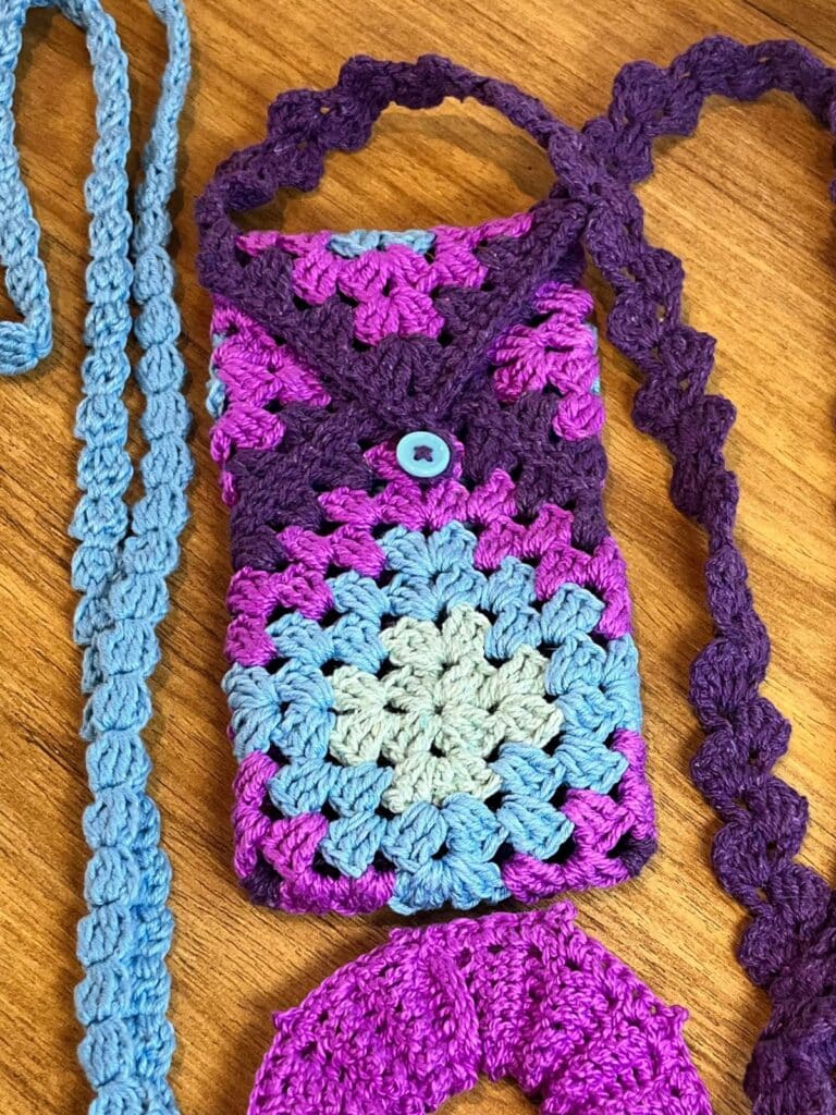 Bolsa para celular de crochê artesanal em tons frios de roxo, lilás e azul claro, com estampa granny square e alça longa trabalhada.