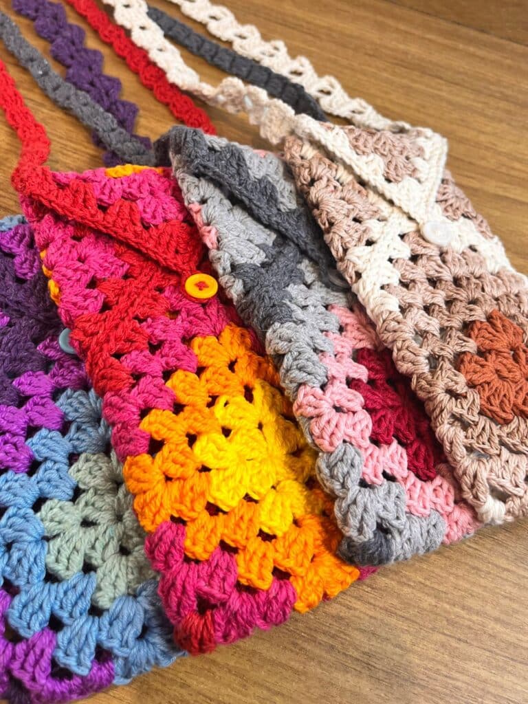 Coleção de bolsas para celular de crochê estilo granny square em diversas paletas de cores, incluindo roxo, vermelho, cinza e tons terrosos, lado a lado sobre uma mesa