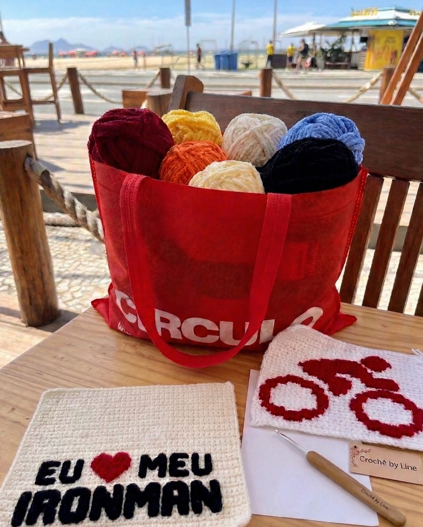 Uma foto de uma sacola vermelha da Círculo cheia de balls de fios pelúcia e aveludados em cores vibrantes, ao lado de um quadrado de crochê com bordado aveludado 'EU ❤️ MEU IRONMAN' e um gráfico impresso.