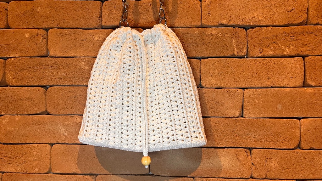 Bolsa de crochê simples e elegante modelo Lara em tom natural suspensa em parede de tijolos rústicos.